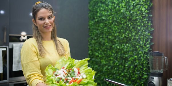 A cheerful woman in a modern kitchen presenting a fresh vegetable salad.